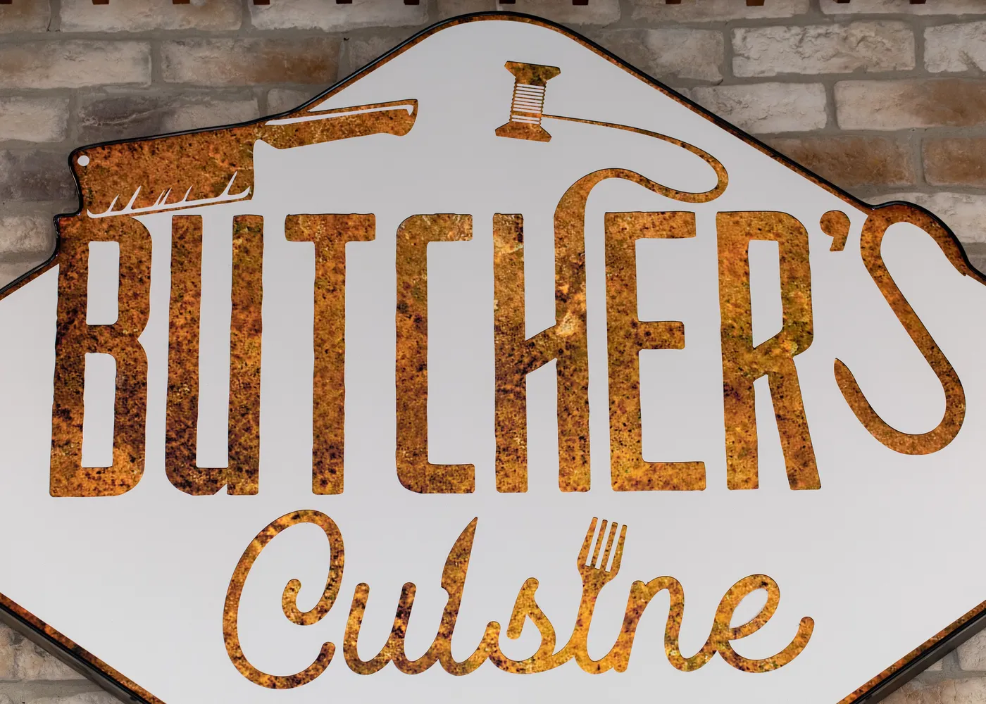 Firma Butcher's Cuisine, litere de metal ruginit pe o placă albă, prinsă pe zidul de cărămidă al magazinului