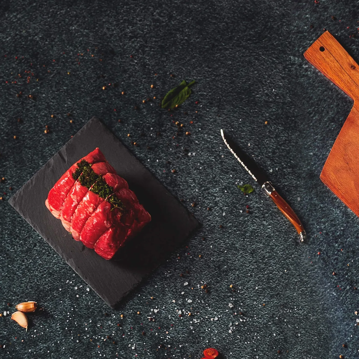 Bucată de roastbeef legată cu sfoară, presărată cu cimbru, pe o ardezie neagră, lângă un cuțit